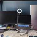 A modern computer workstation with dual monitors, a laptop, keyboard, ring light, and various desk accessories on a white desk against blue walls