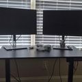 A dual monitor computer setup on a black standing desk positioned in front of windows with white blinds