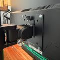 Back view of a desktop computer setup showing a black monitor mounted on a stand, with cables, a black speaker, and a green keyboard visible on a wooden desk surface.