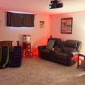 A home theater room with red ambient lighting, featuring brown leather recliners, a projector mounted on the ceiling, blackout curtains, vintage movie posters on the walls, a popcorn machine, and colorful floor cushions on carpeted flooring.