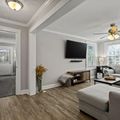 Modern living room with gray sectional sofa, wall-mounted TV, wood flooring, white walls, and ceiling fan with natural light from windows