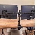 Two Dell computer monitors mounted on a dual monitor arm stand positioned on a wooden desk, with bookshelves visible in the background