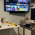 A wall-mounted TV displaying a streaming service menu with various show thumbnails, positioned above a white desk with storage baskets and office supplies in a modern room setup.