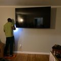 A person in a yellow shirt installing or adjusting a large flat-screen TV mounted on a white wall in a living room with hardwood floors
