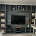 Built-in entertainment center with gray stone fireplace surround, flat screen TV, shelving units with storage baskets, decorative items on mantel, and a small guitar visible on the right side