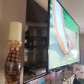 A living room setup with a large wall-mounted TV displaying colorful content, decorative items on a shelf below including a patterned vase with a lamp shade, and various ornamental objects arranged on the entertainment center.