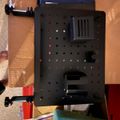 Black pegboard panel with various mounting holes and attachments, positioned on a workbench with tools and materials visible in the background