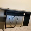 A dark wooden desk with built-in cable management and electrical outlets, positioned against a beige wall with various cables plugged in underneath
