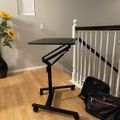 A black adjustable standing desk on wheels positioned near white stair railings, with sunflowers in a vase and a black bag on wooden flooring