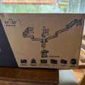 A cardboard box for a dual monitor arm stand that fits 13-32 inch monitors, showing product illustrations and specifications on the packaging