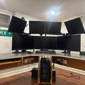 A professional computer workstation with six black monitors arranged in two rows, a keyboard, gaming headphones, and a desktop PC tower on a wooden desk with brown paneling, set in an office space with street signs visible on the walls.