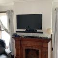 Living room with flat screen TV mounted above wooden fireplace mantel, gray curtains on windows, and partial view of gray couch
