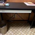 A black desk with white drawer against wood paneling, with stacked papers and a computer mouse on top, cables underneath, and a patterned rug on the floor