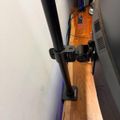 A black microphone stand clamp mounted on a wooden beam or rail, with an orange amplifier visible in the background against a white wall.