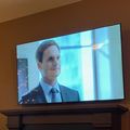 A flat-screen TV mounted on a wall above a wooden mantelpiece, displaying a scene with a man in a business suit and tie in what appears to be an office setting.
