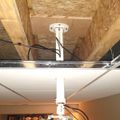 Ceiling-mounted projector installation showing mounting hardware extending through floor joists in an unfinished basement or attic space