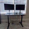 A home office setup with a height-adjustable desk positioned in front of windows with white blinds, featuring two black computer monitors on stands and various cables underneath