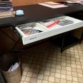 A black desk with an open white drawer containing office supplies, with stacks of papers on top and a waste basket underneath