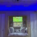 Cozy bedroom nook with built-in seating area featuring pillows and blankets, with a TV mounted above showing a football game, illuminated by blue ambient lighting