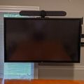 Wall-mounted flat screen TV with video conferencing camera attached on top, positioned near a window with blinds