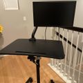 A black adjustable standing desk with a monitor mounted on an arm, positioned next to white stair railings on hardwood flooring