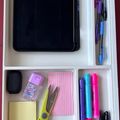 Organized desk drawer with office supplies including a tablet, pens, scissors, sticky notes, highlighters, and other stationery items neatly arranged in compartments