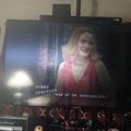Television screen showing Jerry Springer show with blonde woman in red top, with entertainment center and decorative lights below