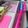 Organized desk drawer containing colorful office supplies including highlighter markers in pink, blue and purple, sticky notes, a stapler, and other stationery items