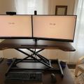 A dual monitor computer setup on an adjustable standing desk with two screens displaying Google homepages, a keyboard and mouse below, and a wooden chair nearby in a home office setting.