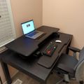 A home office setup with a black standing desk, laptop computer, keyboard, mouse, and office chair in a room with beige walls and window blinds