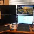A home office setup with multiple monitors, laptops, keyboards and mice arranged on a wooden desk near a window with blinds