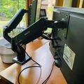A black adjustable monitor arm mounted on a wooden desk holding a computer monitor, with cables visible and a window showing greenery in the background
