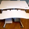 A white adjustable standing desk with two tiers and wheels, positioned on a wooden floor