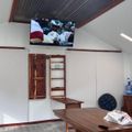 Interior of a room with white walls, wooden trim, vaulted ceiling, wall-mounted TV showing sports content, wooden table with items on it, and a water cooler in the corner