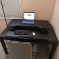 A home office setup with a black standing desk, laptop, keyboard, mouse, and white office chair positioned near a window with blinds