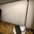 A white desk lamp with an adjustable arm illuminating a smartphone that is wirelessly charging on the lamp's circular base, sitting on a wooden surface