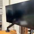A large black Dell computer monitor on a stand positioned on a wooden desk in an office or study area with bookshelves and window blinds visible in the background