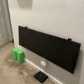 A flat screen monitor mounted on a white wall with green cube-shaped objects and a small black device with cable on the floor below