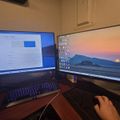 A dual monitor computer setup on a wooden desk showing a Windows settings screen on the left monitor and a scenic sunset desktop wallpaper on the right monitor, with a backlit keyboard and mouse visible