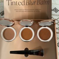 Laura Mercier Tinted Blur Balm makeup display showing three open compacts with different foundation shades and an applicator brush