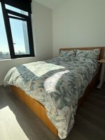 Customer-uploaded image. Click to see more details. The image represents: A modern bedroom with a wooden bed frame dressed in green and cream leaf-patterned bedding. Sunlight streams through a large black-framed window overlooking a forested landscape. The room features light beige walls and dark flooring.