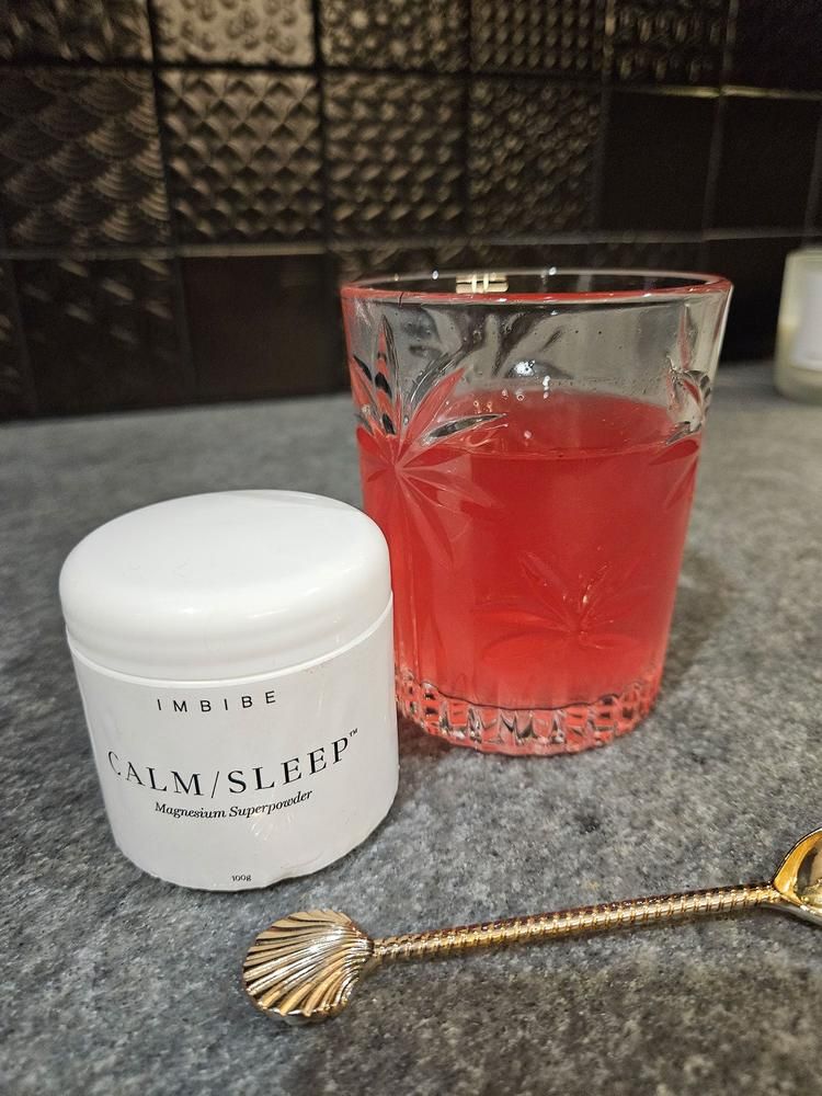 A CALM/SLEEP jar beside a glass of red drink and a gold spoon on a granite counter