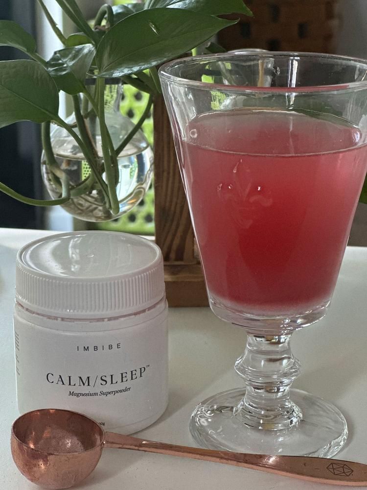 A glass of pink drink beside a CALM/SLEEP jar, copper scoop and plant
