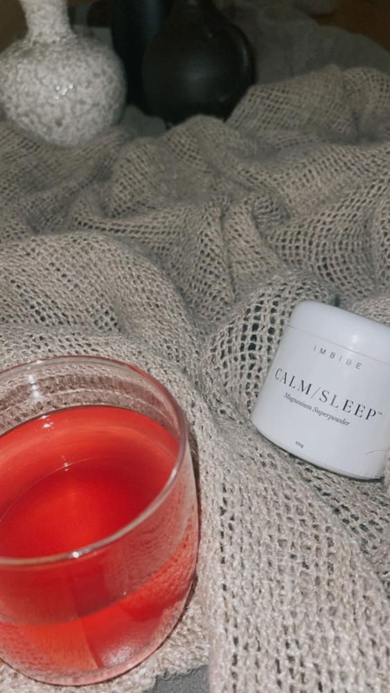 A glass of red drink beside a CALM/SLEEP jar on textured beige fabric