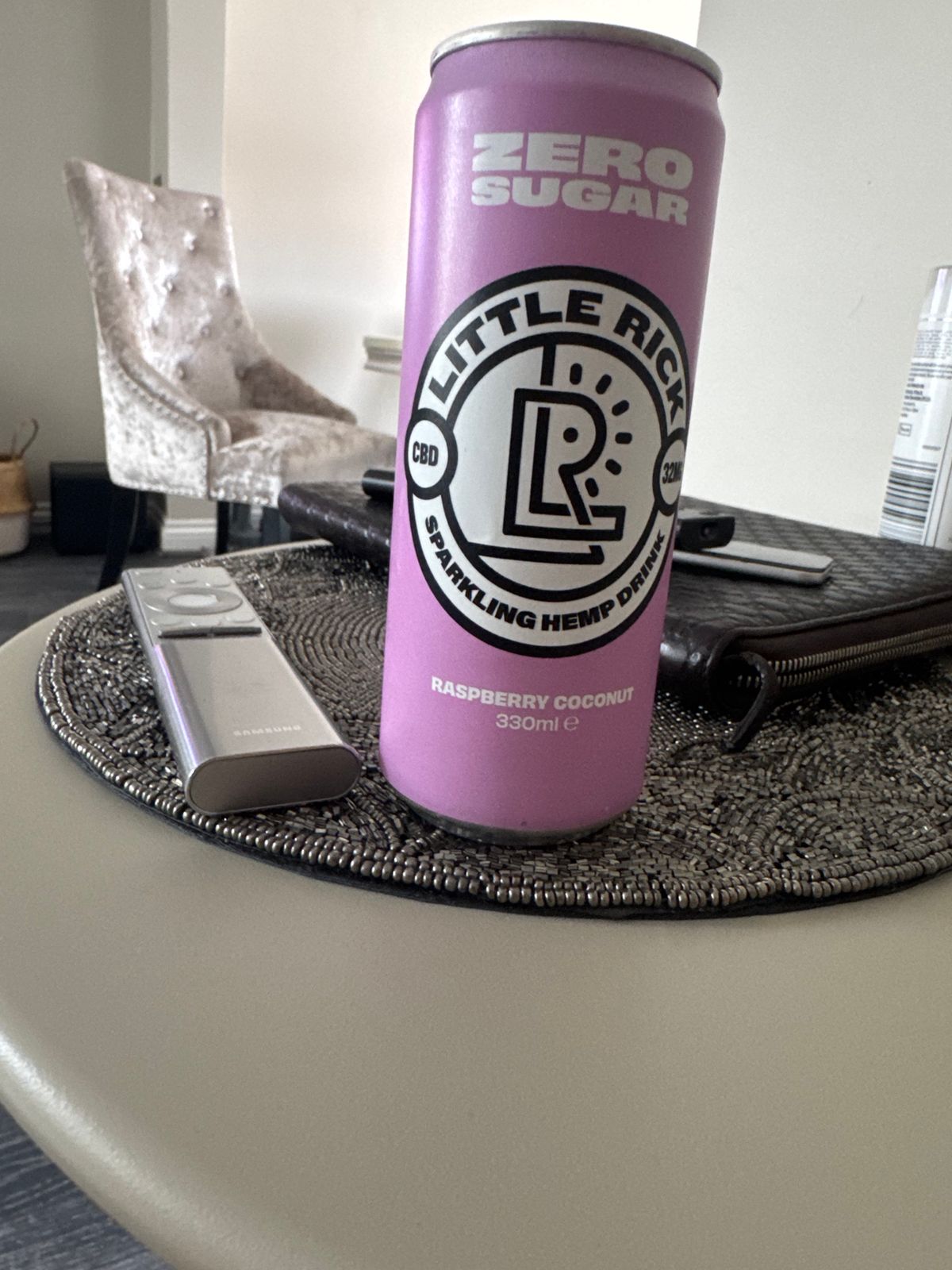A pink can of Little Rick CBD sparkling hemp drink in raspberry coconut flavor sits on a woven placemat next to a phone on a white table