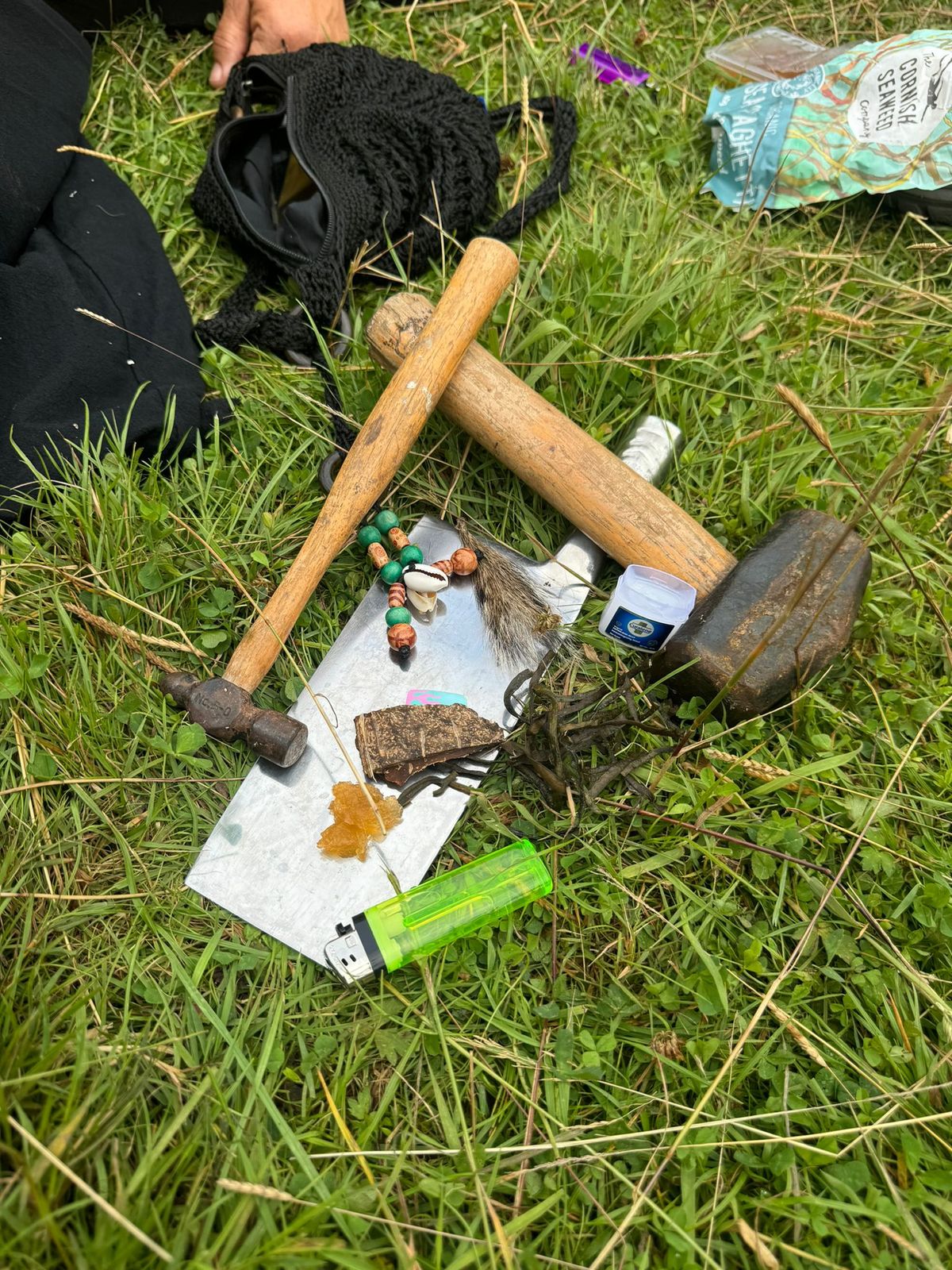 Traditional flint knapping tools and materials laid out on grass including wooden-handled hammers, stone pieces, and various crafting supplies