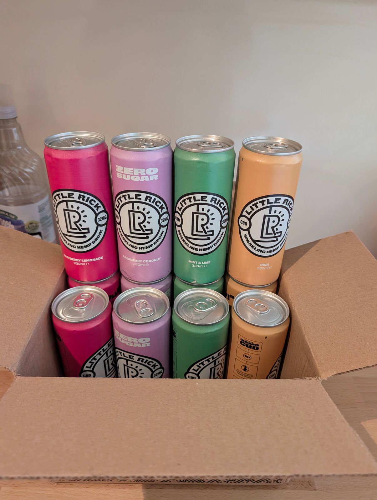 Eight colorful Little Ricey sparkling hemp drink cans in a cardboard box, featuring pink, purple, green, and orange varieties
