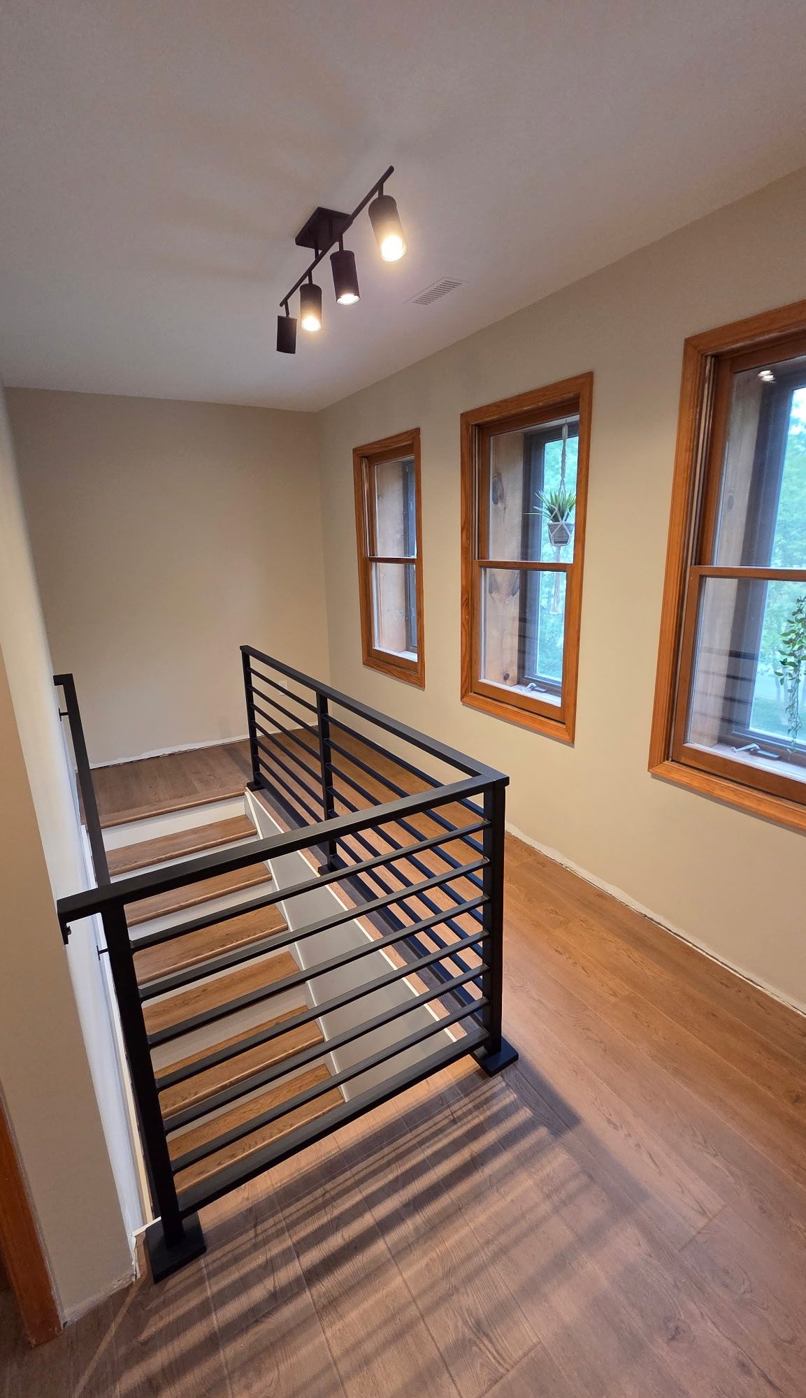 Interior stairway landing with black metal railing, wood floors, three windows with wood trim, and track lighting on ceiling