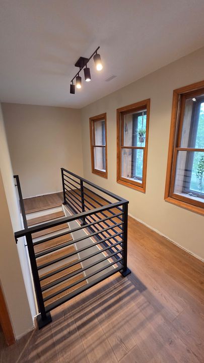 Interior stairway landing with black metal railing, wood floors, three windows with wood trim, and track lighting on ceiling