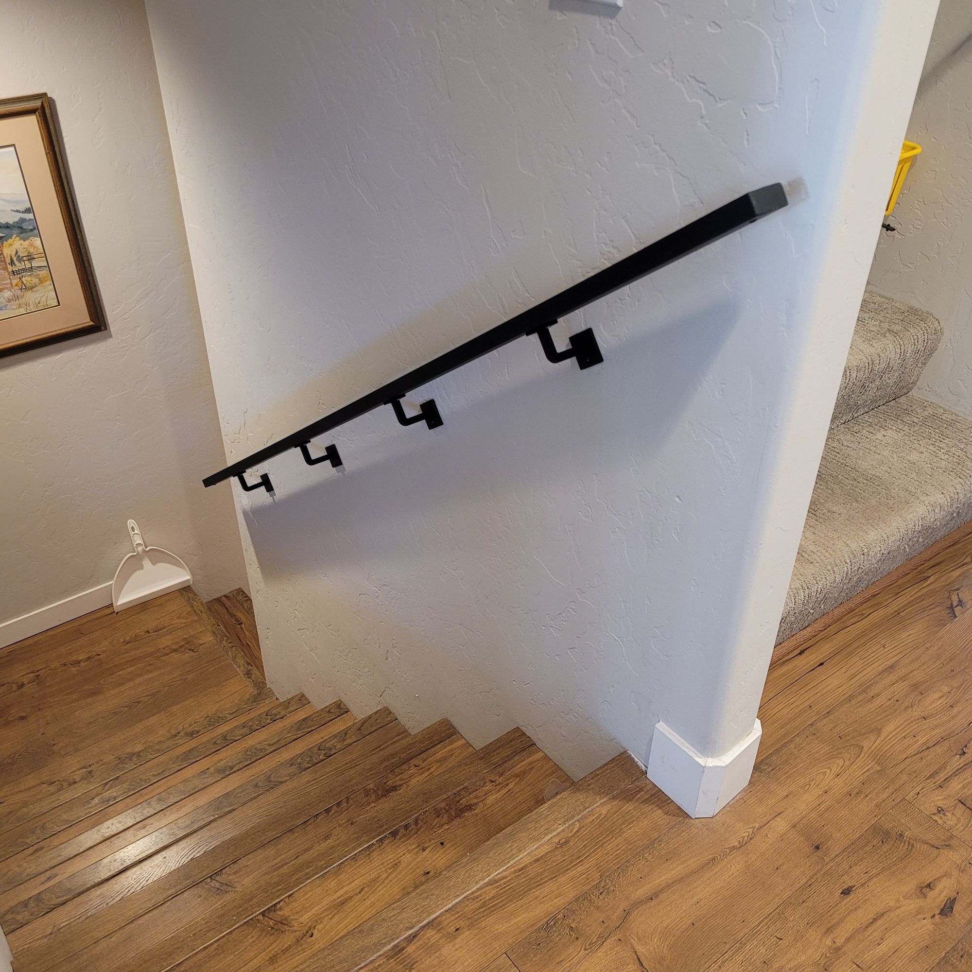 Interior staircase with black metal handrail mounted on white wall, wooden flooring, and carpeted steps visible
